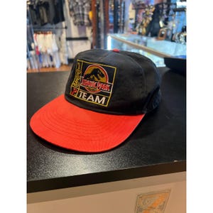 Può includere: Un cappellino da baseball nero con visiera rossa caratterizzato da una toppa ricamata sul davanti. La toppa mostra la sagoma di un dinosauro, il testo Jurassic Park, l'anno 1993 e la parola Team in lettere gialle, rosse e bianche su sfondo nero.