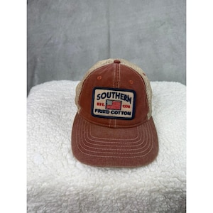 Può includere: Cappellino da camionista rosso e beige con una toppa che recita "SOUTHERN EST. 1776 FRIED COTTON" con una bandiera americana. Il cappello ha una visiera curva e un retro in rete.