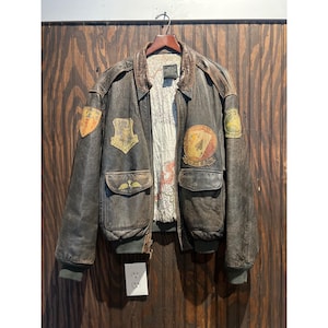 Puede incluir: Una chaqueta bomber de cuero marrón oscuro con varios parches bordados en el pecho y las mangas, incluido un diseño de cartas de juego. El forro interior muestra un mapa impreso, y la chaqueta tiene puños y cuello acanalados.