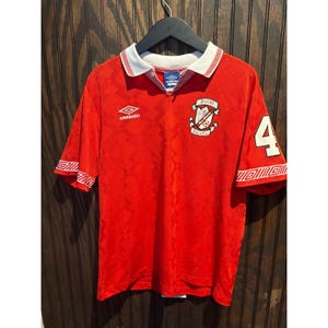 May include: A red short-sleeve soccer jersey featuring a white collar with red trim, an Umbro logo on the right chest, and a Point Soccer crest on the left. The number 4 is printed in white on the left sleeve, and the fabric has a subtle geometric pattern.