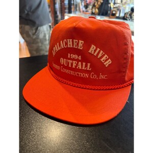 Può includere: Un cappellino da baseball arancione brillante con un bordo in corda intrecciata alla base della corona. La parte anteriore presenta un testo ricamato di colore chiaro che recita APALACHEE RIVER 1994 OUTFALL SHERIFF CONSTRUCTION CO., INC. in un design strutturato.