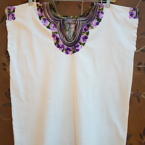 May include: White sleeveless blouse with a purple and green embroidered floral design around the neckline.