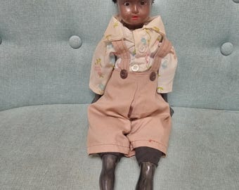 May include: A vintage black doll with a brown and white patterned shirt and brown overalls. The doll has black hair and a painted face.