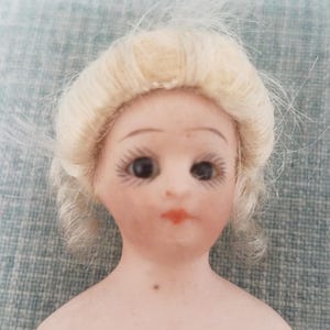 May include: A porcelain doll head with blonde hair styled in a bun. The doll has painted eyes, eyebrows, and lips. The doll's neck is visible, and there is a small brown dot on the doll's chest.