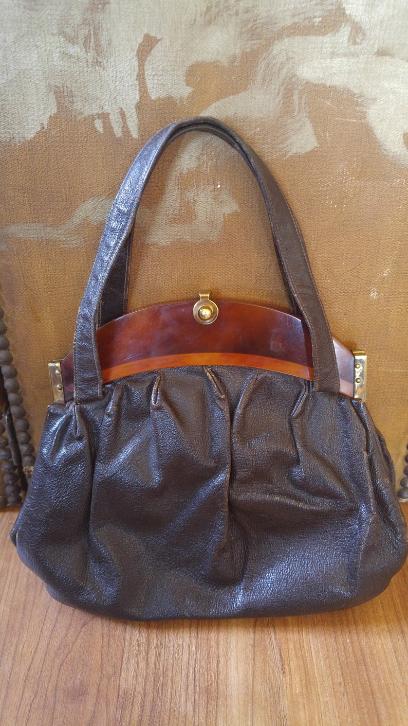 Puede incluir: Un bolso de mano de cuero marr&oacute;n con un asa superior de pl&aacute;stico marr&oacute;n y detalles dorados.