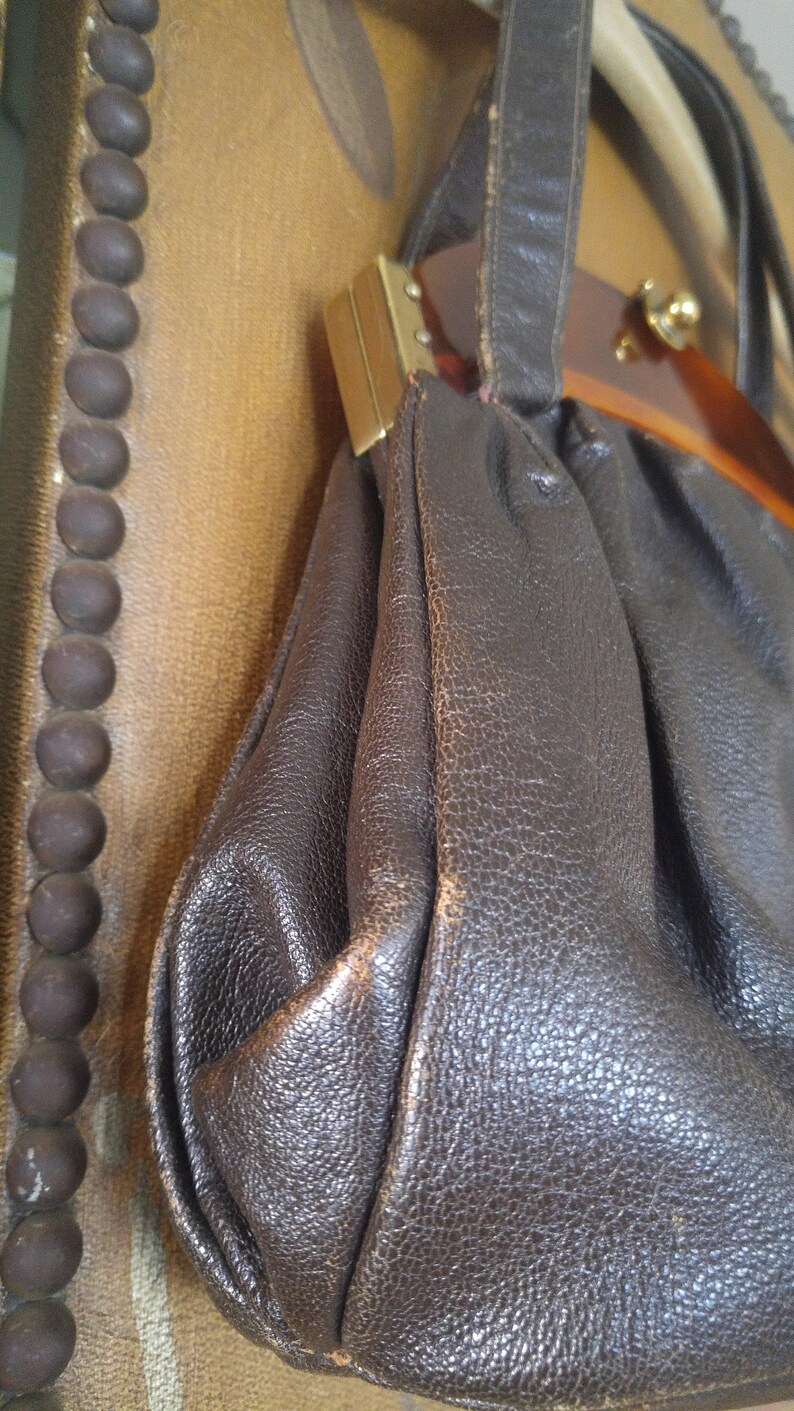 Puede incluir: Un bolso de mano de cuero marr&oacute;n con asa superior y cierre dorado. El bolso tiene una forma redondeada y un exterior de cuero texturizado.