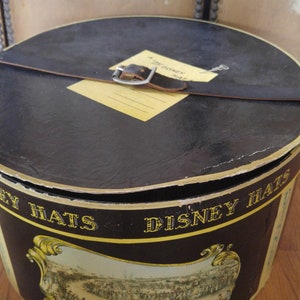 May include: A black hat box with a leather strap and buckle. The box has a yellow label that says "A Disney Hat". The box is decorated with a scene of people at a horse race. The text "Disney Hats" is printed on the side of the box.