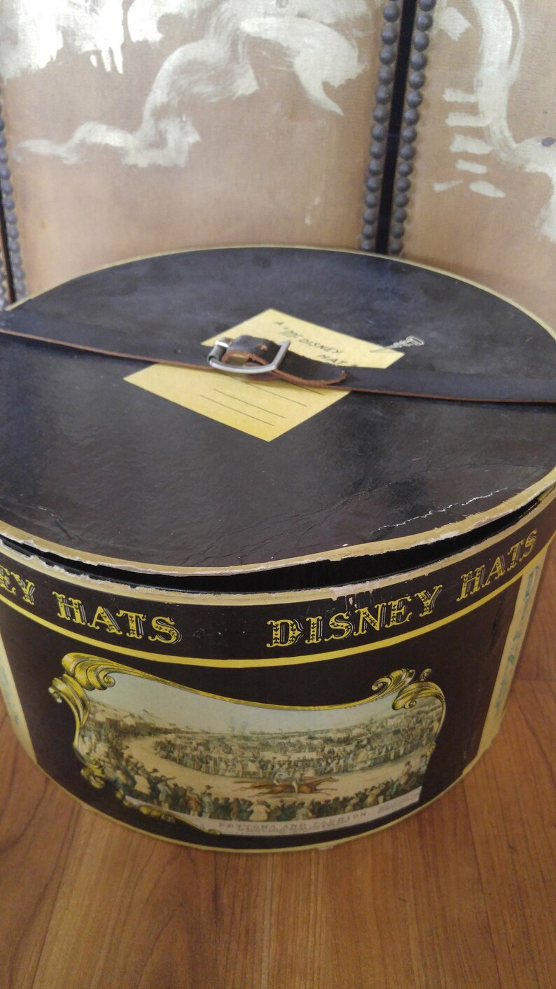 May include: A black hat box with a leather strap and buckle. The box has a yellow label that says "A Disney Hat". The box is decorated with a scene of people at a horse race. The text "Disney Hats" is printed on the side of the box.