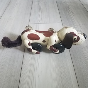 May include: A vintage tin toy dog with brown and white spots. The dog has black wheels and a brown felt tail. The toy is on a light wood floor.