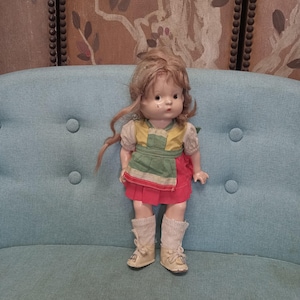 May include: A vintage porcelain doll with blonde hair, wearing a green and red dress and white socks with brown shoes. The doll is sitting on a blue couch.