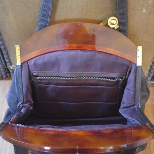 Puede incluir: Un bolso de mano vintage de cuero negro con una parte superior de pl&aacute;stico marr&oacute;n y una cremallera dorada. El bolso tiene una sola correa y un cierre de solapa.