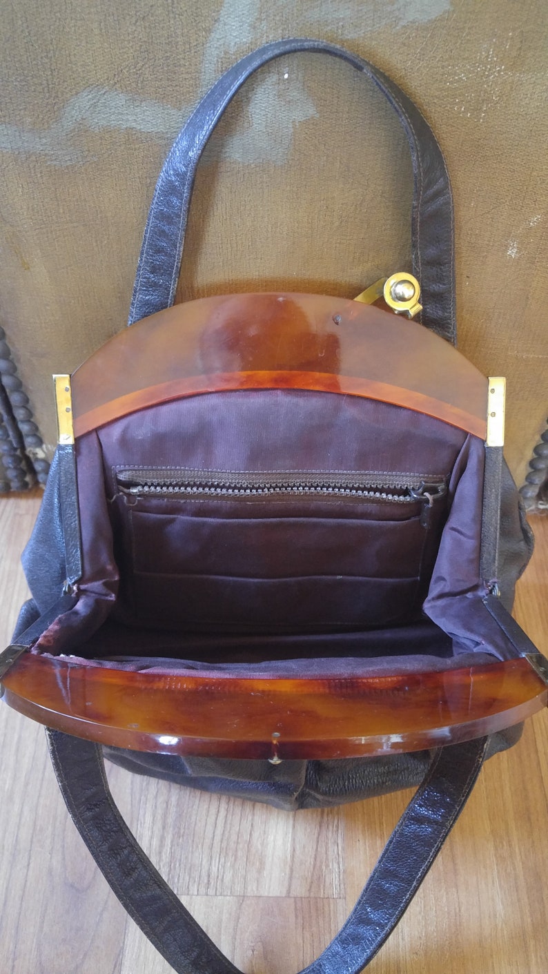 Puede incluir: Un bolso de mano vintage de cuero negro con una parte superior de pl&aacute;stico marr&oacute;n y una cremallera dorada. El bolso tiene una sola correa y un cierre de solapa.