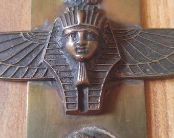 May include: A brass pendant necklace with three decorative elements: a lion's head, a winged Egyptian pharaoh, and a profile of a woman with a floral design. The pendant is attached to a chain.