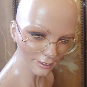 May include: A close-up of a mannequin's face wearing gold-rimmed glasses. The mannequin has a smooth, pale complexion and is wearing a neutral expression.
