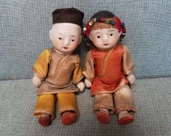 May include: Two antique porcelain dolls, a boy and a girl, are seated on a blue upholstered surface. The boy doll wears a brown hat, gold and brown clothing, and red shoes. The girl doll has a beaded headband, red and tan clothing, and red shoes.