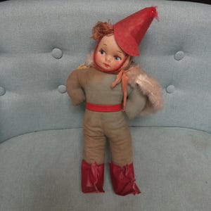 May include: A vintage doll with a red hat and boots, wearing a green and brown outfit. The doll is sitting on a light blue chair.