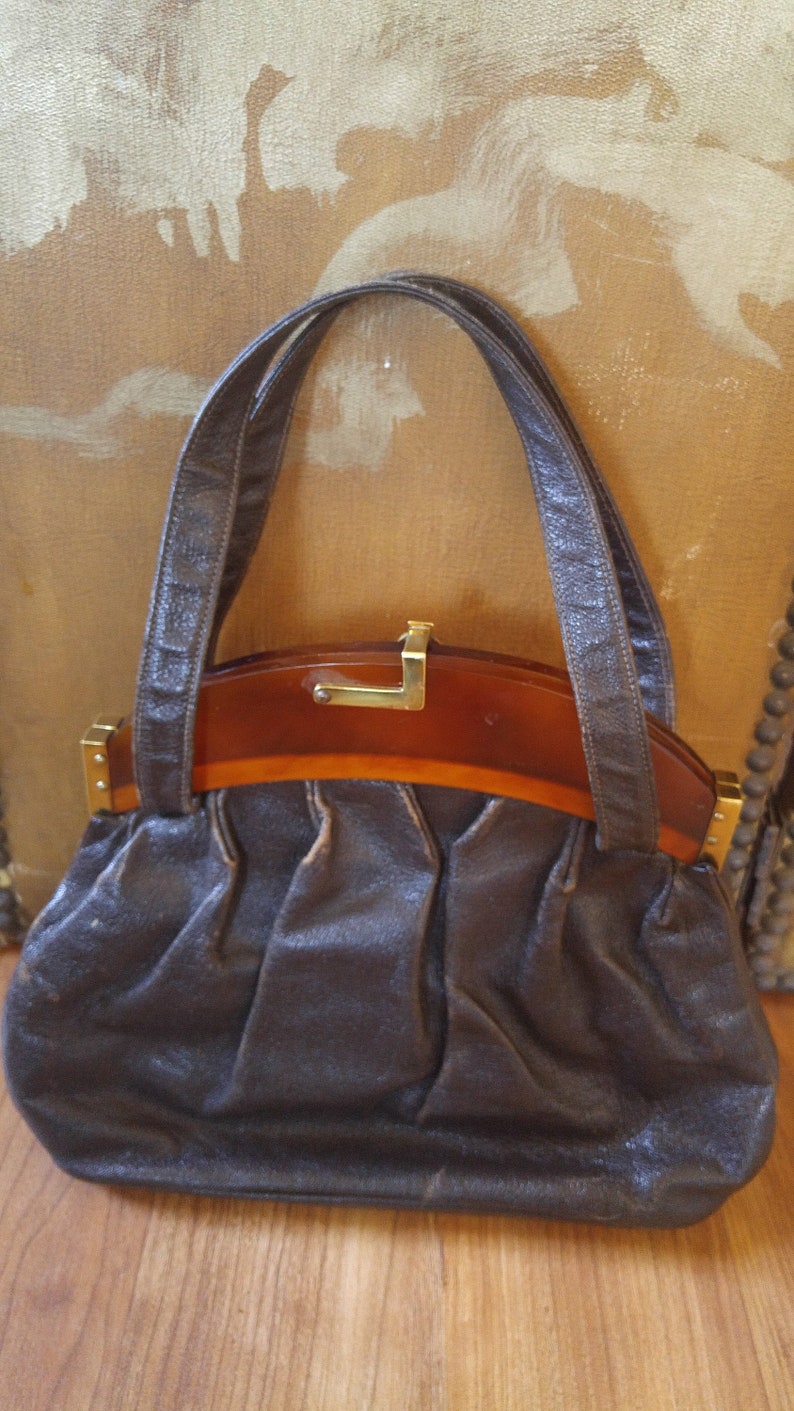 Puede incluir: Un bolso de mano de cuero marr&oacute;n con un asa superior de carey y detalles dorados.