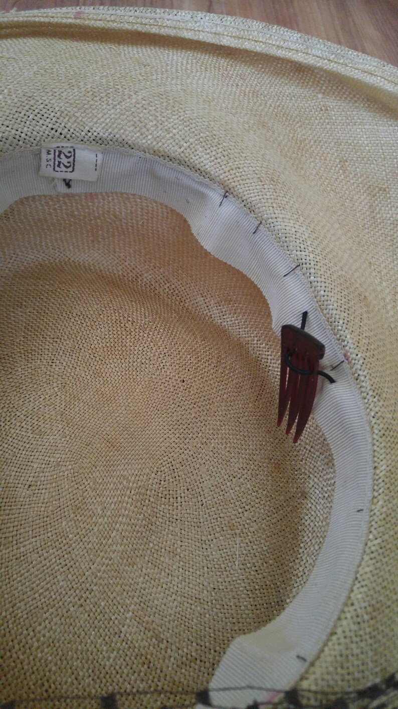 May include: The inside of a straw hat with a white ribbon and a brown comb attached to the ribbon. The hat is made of woven straw and has a label that says "22" on it.