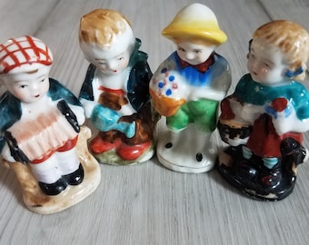 Puede incluir: Cuatro figuritas de porcelana de niños, cada una vestida con ropa diferente y sosteniendo varios objetos. Las figuritas están pintadas en colores brillantes, incluyendo rojo, azul, verde, amarillo y blanco.