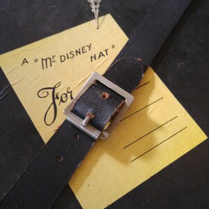 May include: A yellow card with the text "A "M" DISNEY HAT" and the word "For" written in cursive. A black leather strap with a silver buckle is attached to the card.