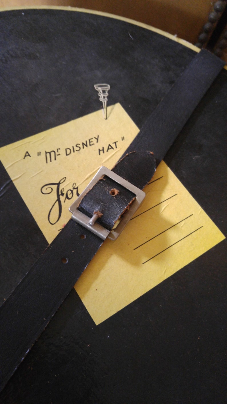 May include: A yellow card with the text "A "M" DISNEY HAT" and the word "For" written in cursive. A black leather strap with a silver buckle is attached to the card.