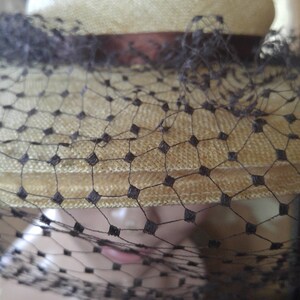 May include: A straw hat with a black veil. The hat is a light beige color and has a wide brim. The veil is made of a fine mesh and covers the entire hat.