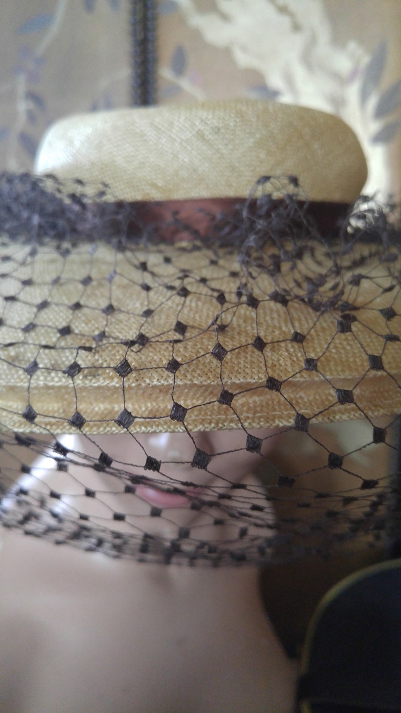 May include: A straw hat with a black veil. The hat is a light beige color and has a wide brim. The veil is made of a fine mesh and covers the entire hat.