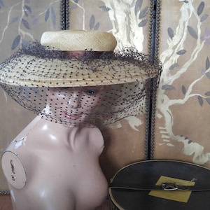 May include: A straw hat with a black veil on a mannequin. The hat is sitting on a table with a black and yellow hat box that says "Disney Hats" on it.