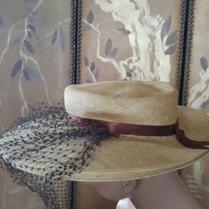 May include: A straw hat with a brown ribbon and a black netting veil. The hat is on a mannequin's head.