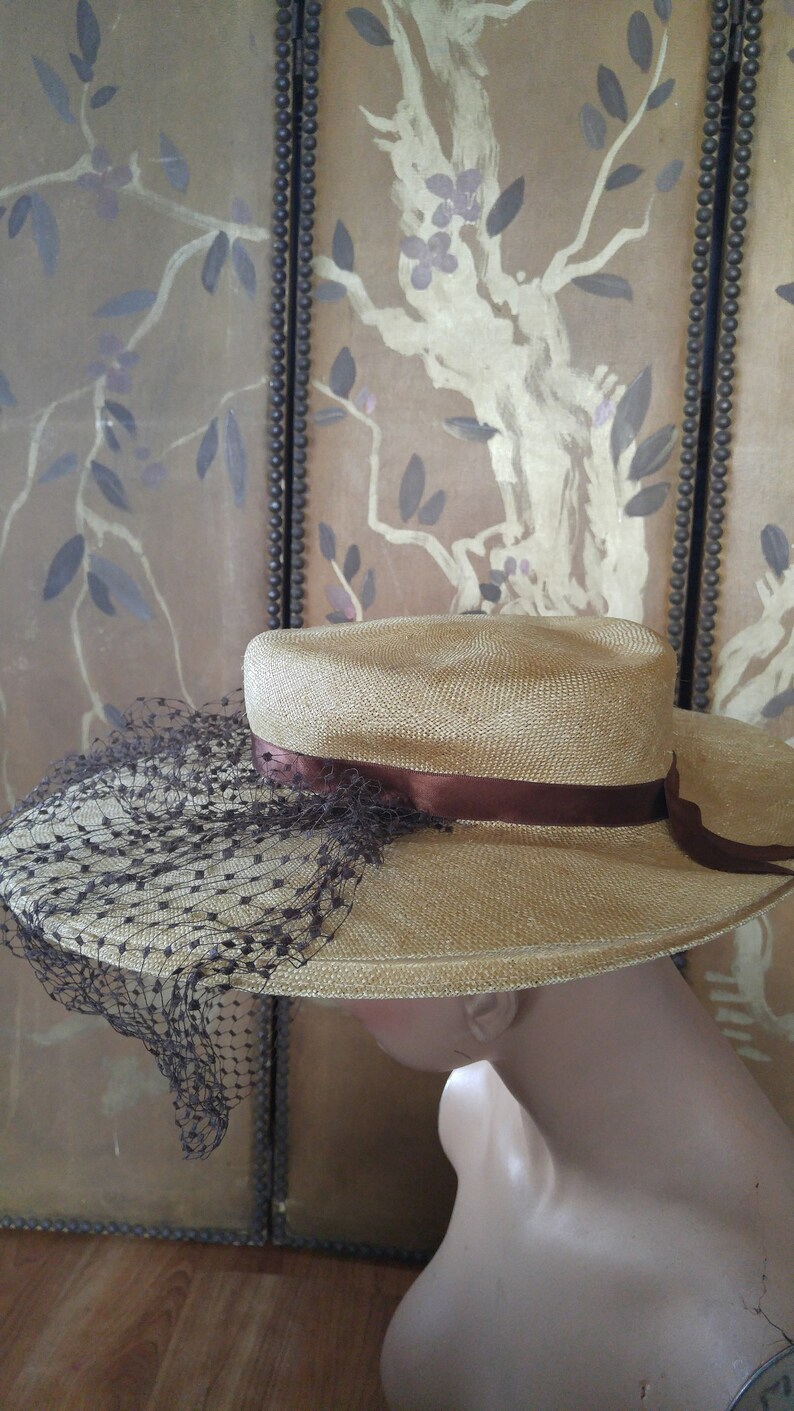 May include: A straw hat with a brown ribbon and a black netting veil. The hat is on a mannequin's head.