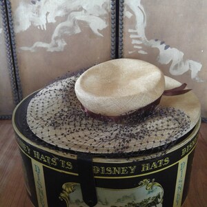 May include: A vintage hat box with a black and gold design featuring a horse-drawn carriage scene. The box is labeled "Disney Hats" and has a brown hat with a black net veil inside.