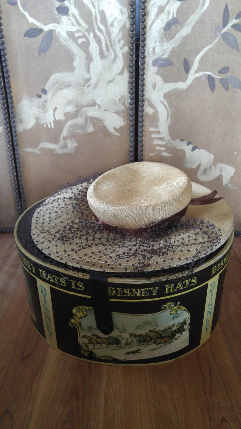 May include: A vintage hat box with a black and gold design featuring a horse-drawn carriage scene. The box is labeled "Disney Hats" and has a brown hat with a black net veil inside.