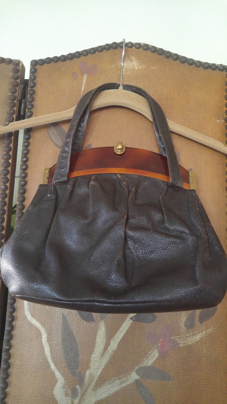 Puede incluir: Un bolso de mano de cuero marr&oacute;n con un asa superior de carey y detalles dorados.