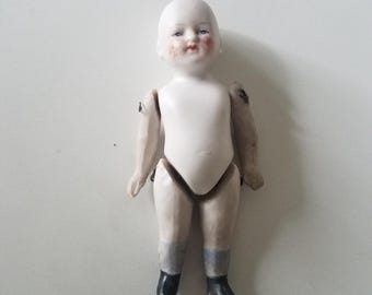 May include: A vintage porcelain doll with a bisque finish. The doll has a white torso, jointed arms and legs, and painted facial features including rosy cheeks and blue eyes. The doll is wearing blue socks and black shoes.