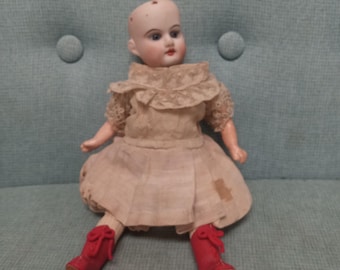 May include: A vintage porcelain doll with a bald head and painted features. The doll is wearing a white dress with lace trim and red shoes.