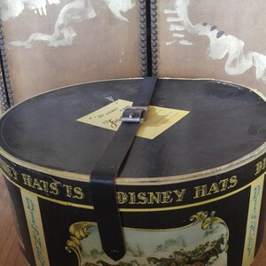 May include: A black hat box with a gold and black label that says "Disney Hats" and features a scene of horses pulling a carriage. The box has a leather strap with a metal buckle.