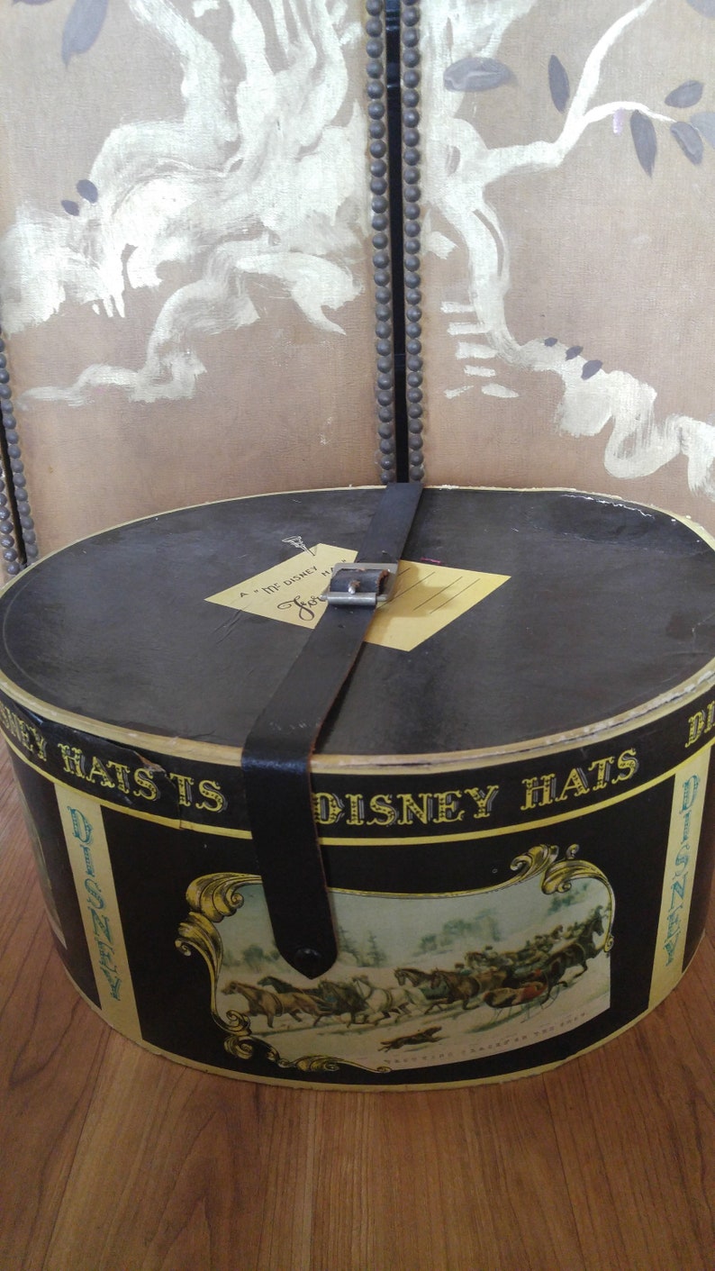 May include: A black hat box with a gold and black label that says "Disney Hats" and features a scene of horses pulling a carriage. The box has a leather strap with a metal buckle.