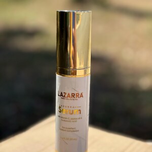May include: A cylindrical serum bottle with a shiny gold cap and base. The white label features the text LAZARRA, Serum, with vitamin C, jojoba oil & hyaluronic acid, and 1.4 FL OZ (35 mL) printed in black and gold lettering on the front surface.