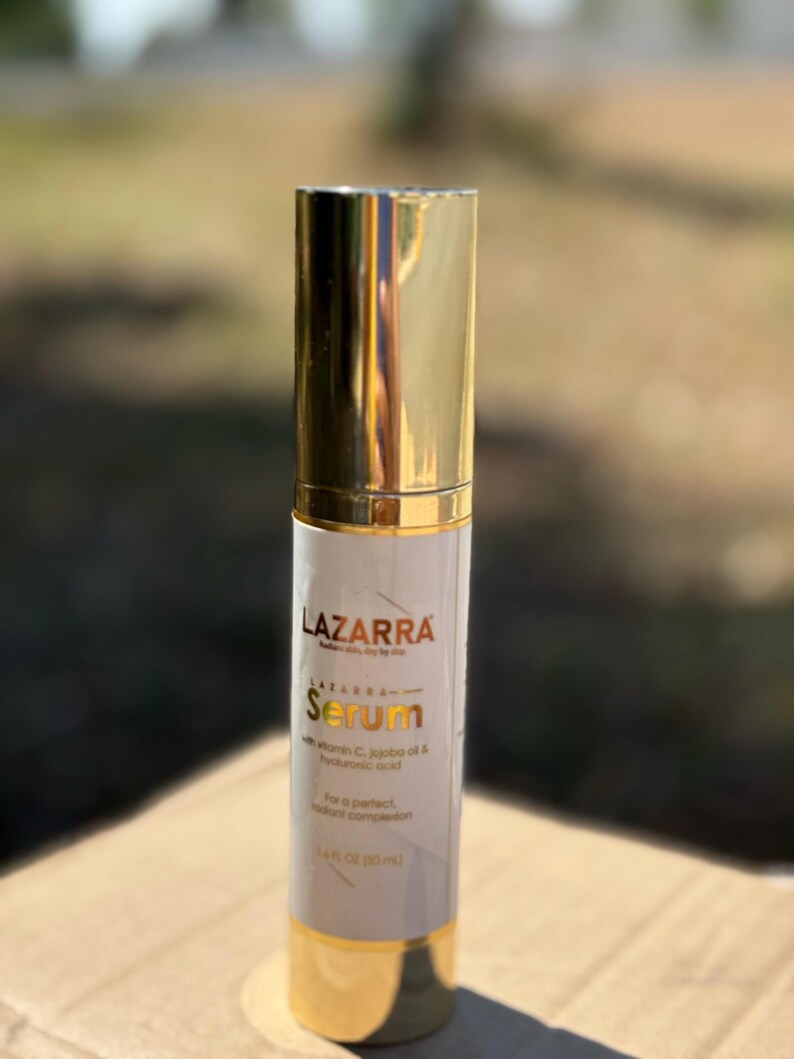 May include: A cylindrical serum bottle with a shiny gold cap and base. The white label features the text LAZARRA, Serum, with vitamin C, jojoba oil & hyaluronic acid, and 1.4 FL OZ (35 mL) printed in black and gold lettering on the front surface.