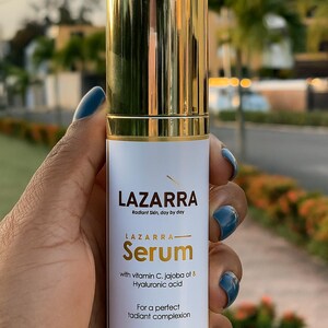 May include: A cylindrical serum bottle with a reflective gold cap and base. The white label features the text LAZARRA, Radiant Skin, day by day, LAZARRA Serum, with vitamin C, jojoba oil & Hyaluronic acid, For a perfect radiant complexion, 1.6 FL OZ (50 mL).