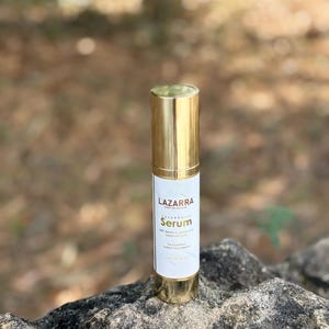 May include: A cylindrical bottle of Lazzara Serum with a metallic gold cap and base. The white label features the text Lazzara Serum with vitamin C, vitamin E & hyaluronic acid, for a perfect radiant complexion, and 1.0 FL OZ (30 ml).
