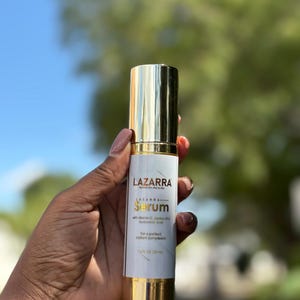 May include: A cylindrical bottle of LAZARRA serum with a gold cap and base. The white label features the brand name, the word Serum, and lists ingredients including vitamin C, jojoba oil, and hyaluronic acid, with a volume of 1.6 fl oz.