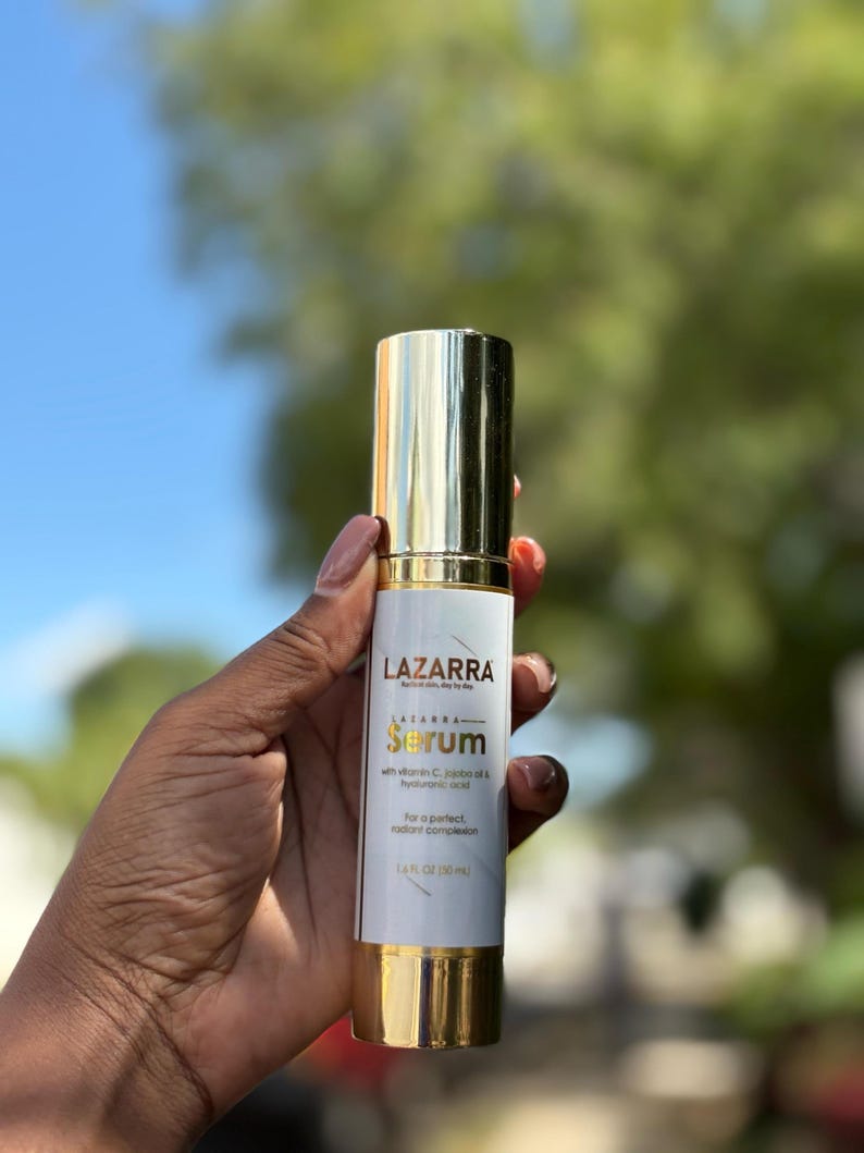 May include: A cylindrical bottle of LAZARRA serum with a gold cap and base. The white label features the brand name, the word Serum, and lists ingredients including vitamin C, jojoba oil, and hyaluronic acid, with a volume of 1.6 fl oz.