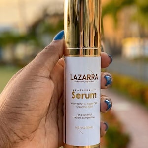 May include: A cylindrical bottle of LAZARRA serum with a shiny gold cap and base. The white label features the text LAZARRA, Radiant skin, day by day, Serum, With Vitamin C, Jojoba Oil & Hyaluronic Acid, For a powerful radiant complexion, 1.69 FL OZ (50mL).