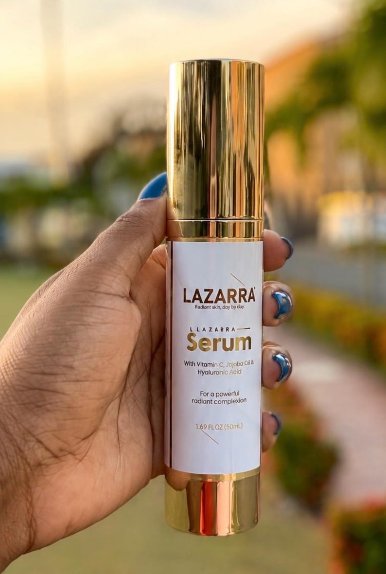 May include: A cylindrical bottle of LAZARRA serum with a shiny gold cap and base. The white label features the text LAZARRA, Radiant skin, day by day, Serum, With Vitamin C, Jojoba Oil & Hyaluronic Acid, For a powerful radiant complexion, 1.69 FL OZ (50mL).
