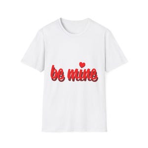 Peut inclure: T-shirt blanc avec l'inscription "be mine" en rouge, en écriture cursive. Un petit cœur rouge est au-dessus du mot "mine". Le t-shirt est de style col rond.