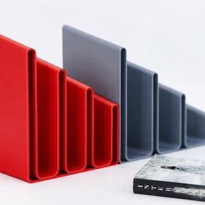 May include: Red and grey modern magazine holders with a stepped design. The red holder has four sections, and the grey holder has five. A book with a grey cover is in front of the holders.