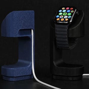 Op de afbeelding: Twee horloge oplaadstandaards, één marineblauw en één zwart, met een witte oplaadkabel. De zwarte standaard houdt een smartwatch met een zwarte band en een kleurrijk display vast. Beide standaards hebben een getextureerd, geribbeld ontwerp.