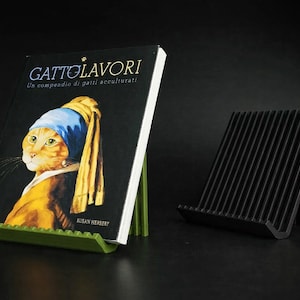 Può includere: Un libro intitolato "GATTO LAVORI" con un ritratto di gatto nello stile de "La ragazza con l'orecchino di perla" di Vermeer è esposto su un leggio verde. Un leggio nero a doghe è sulla destra. Lo sfondo è nero.