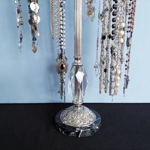 May include: A silver-toned jewelry display stand with a marble base and a faceted crystal accent. Various necklaces of different styles and lengths hang from the stand, showcasing a variety of beads, charms, and pendants. The background is a light blue.